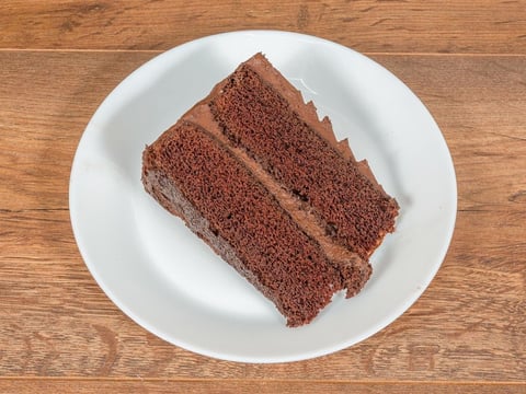 Slice of layered chocolate cake on a white plate, placed on a wooden surface