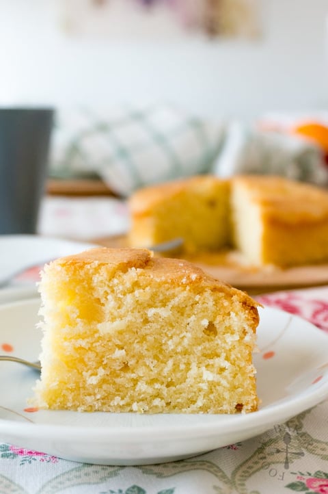 Slice of golden yellow butter cake on white plate with whole cake in background on cutting board