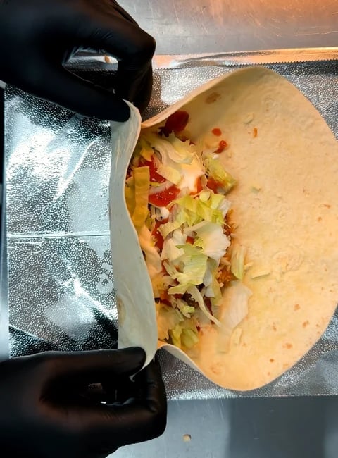 Gloved hands holding an open flour tortilla filled with lettuce, diced tomatoes, and other taco ingredients