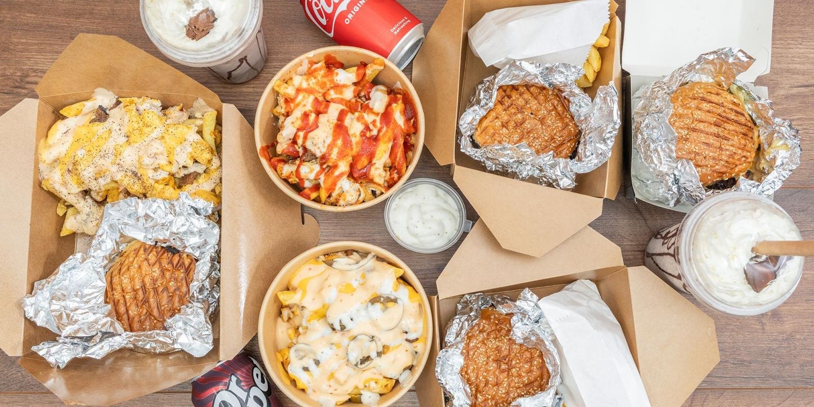 Overhead view of fast food meal spread including fried chicken sandwiches, waffle fries, side dishes, and beverages on wooden table
