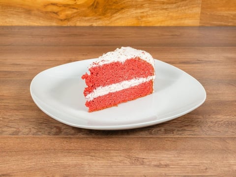 Slice of red velvet cake with white frosting on a white plate on a wooden surface