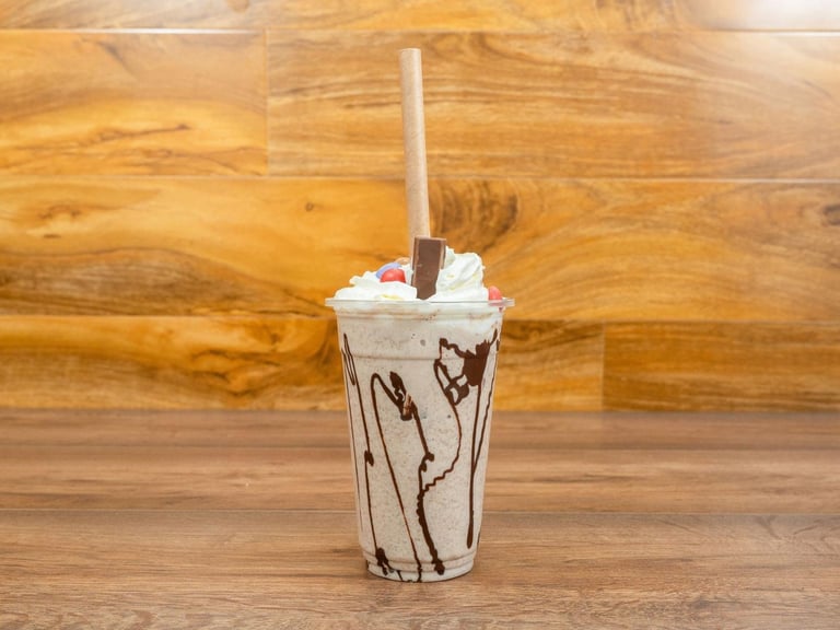Iced beverage in clear cup with chocolate drizzle, topped with whipped cream and a chocolate stick, served on wooden surface